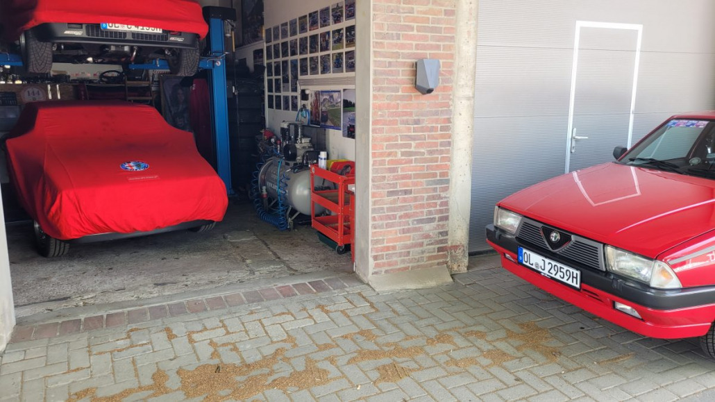 Eine Autogarage mit einer roten Autoabdeckung auf einem Auto, einer davor geparkten roten Alfa Romeo Limousine und verschiedenen Werkzeugen und Geräten entlang der Wand.